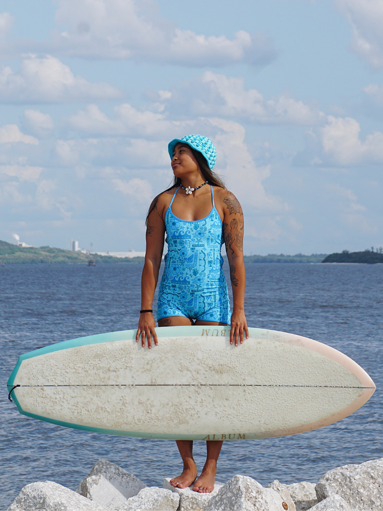 Women holding a surfboard by the water in a blue patterned shorts style surf suit one piece