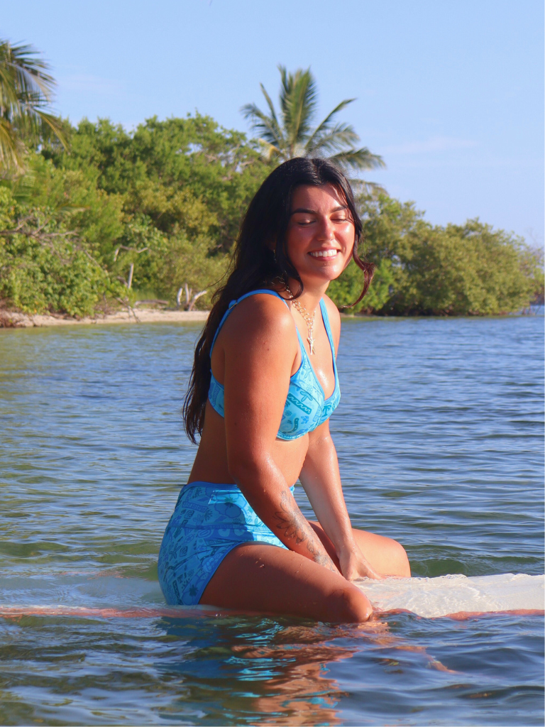 Woman in a blue surf bikini sitting in water with trees and sky in the background