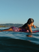 Person lying on a surfboard in the water with a scenic background wearing a pink and emerald green one piece open back shorts style surf suit